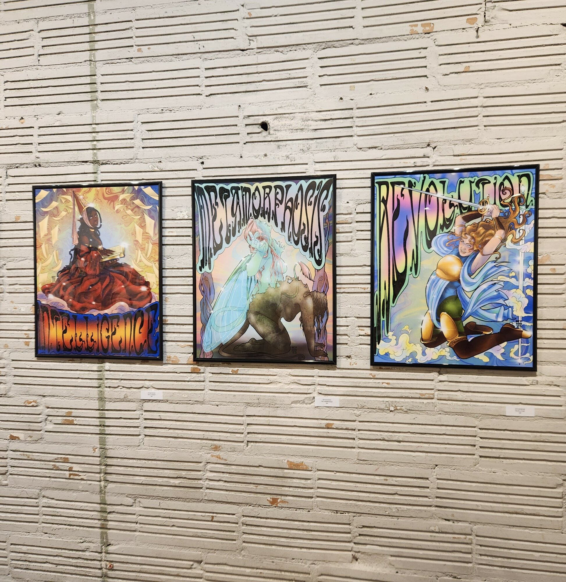 Three framed posters on a wooden wall, featuring abstract and colorful designs with figures and text.