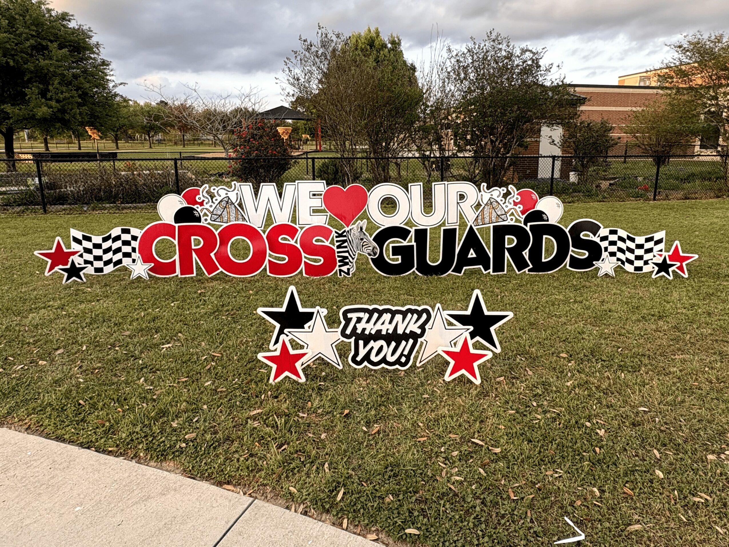 Decorative sign with text and stars on grass, trees, and a building in background, celebrating cross guards.