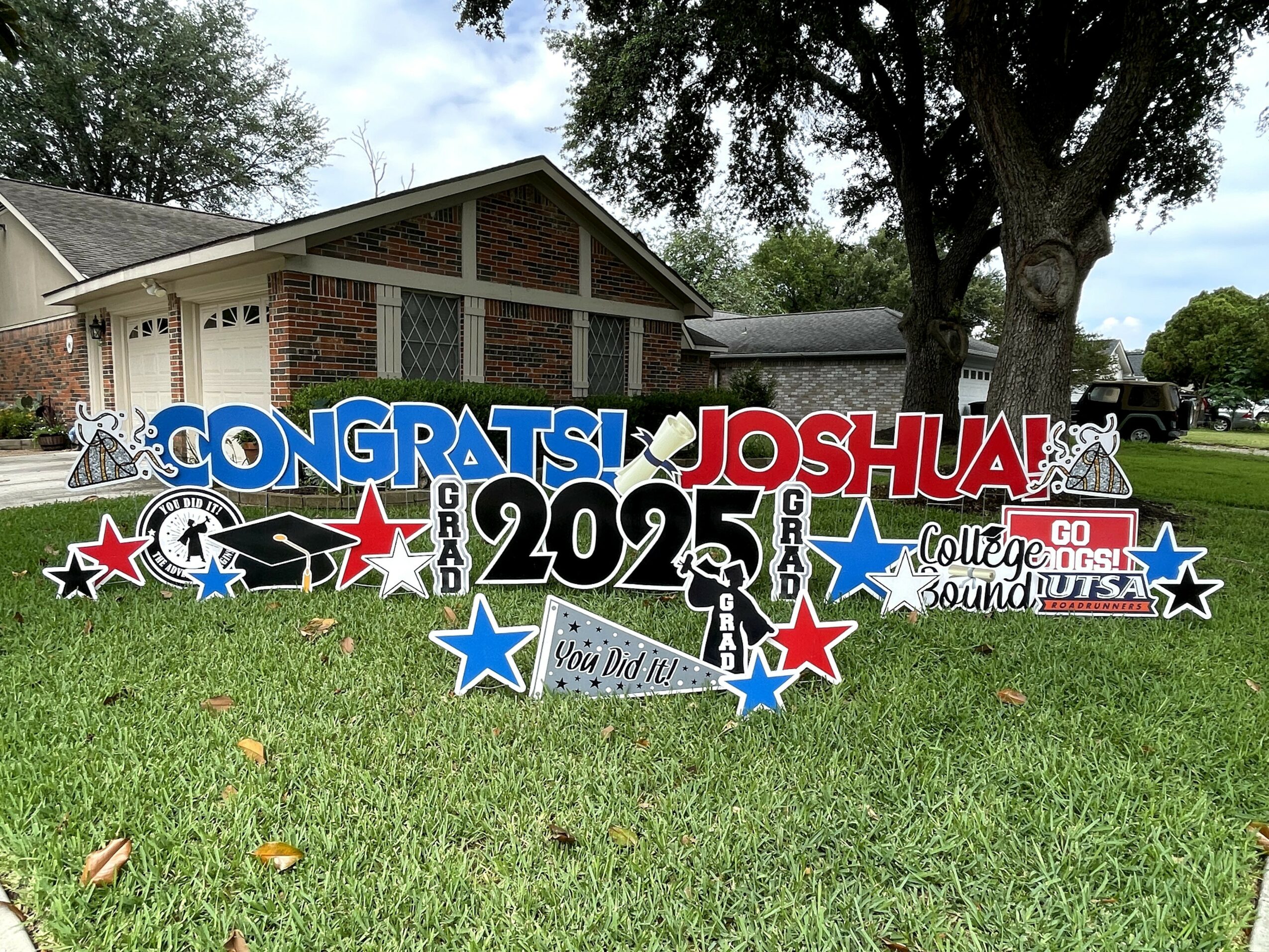 Graduation yard sign with congratulatory messages, stars, and a person in front, on a grassy lawn with trees and houses.