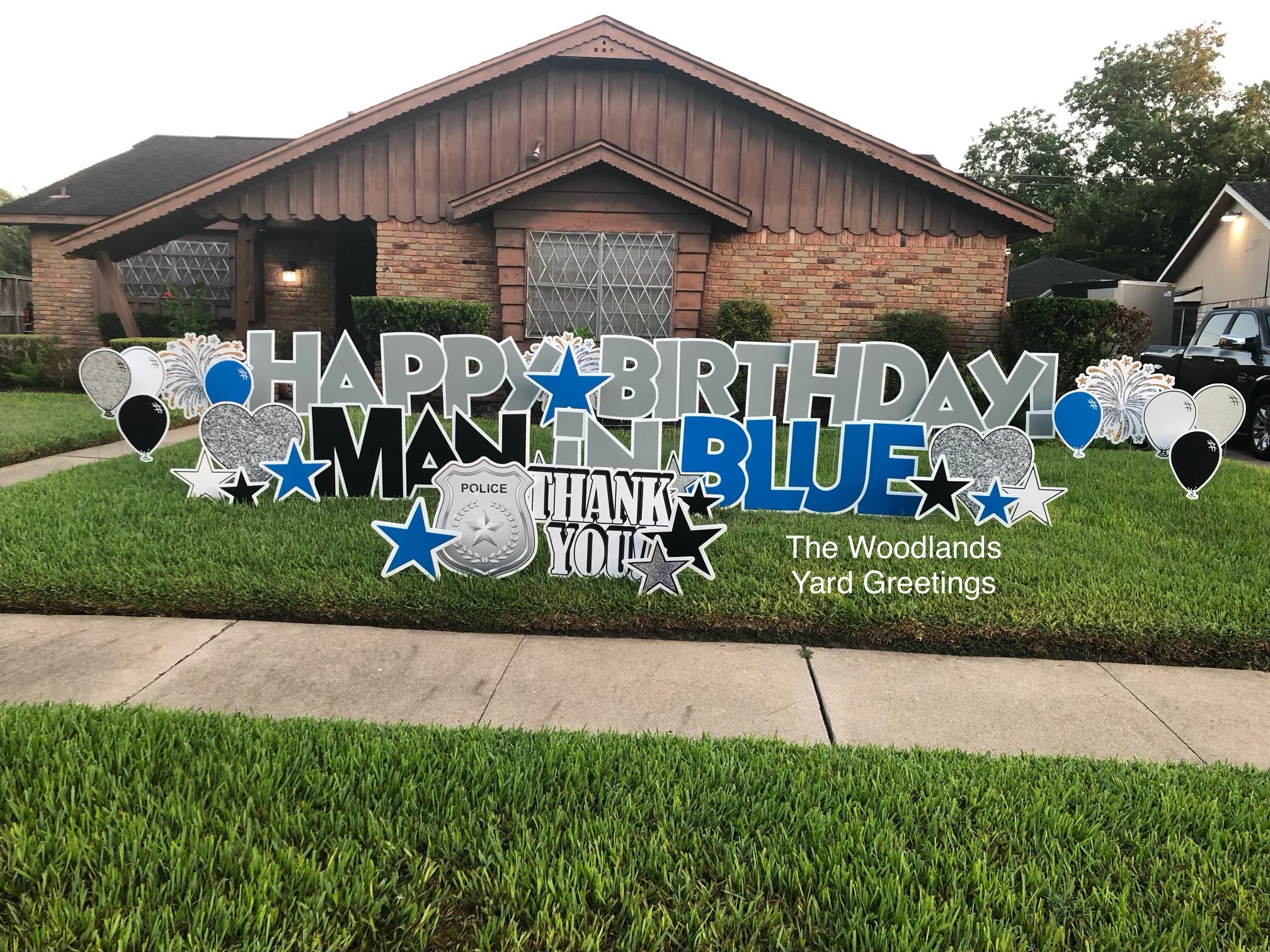 Front yard with birthday decorations, including balloons and signs, in front of a house with a brown roof and brick exterior.