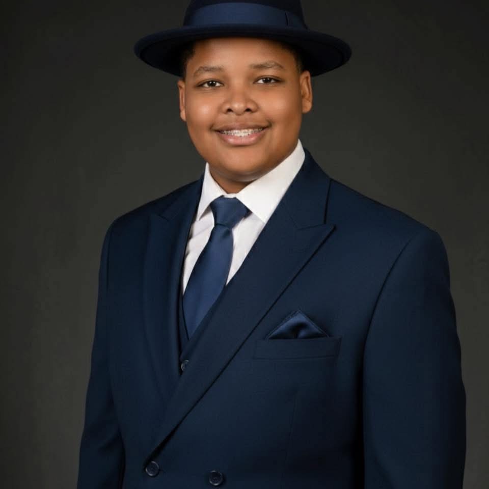 Person in formal suit and hat smiling, dark background.