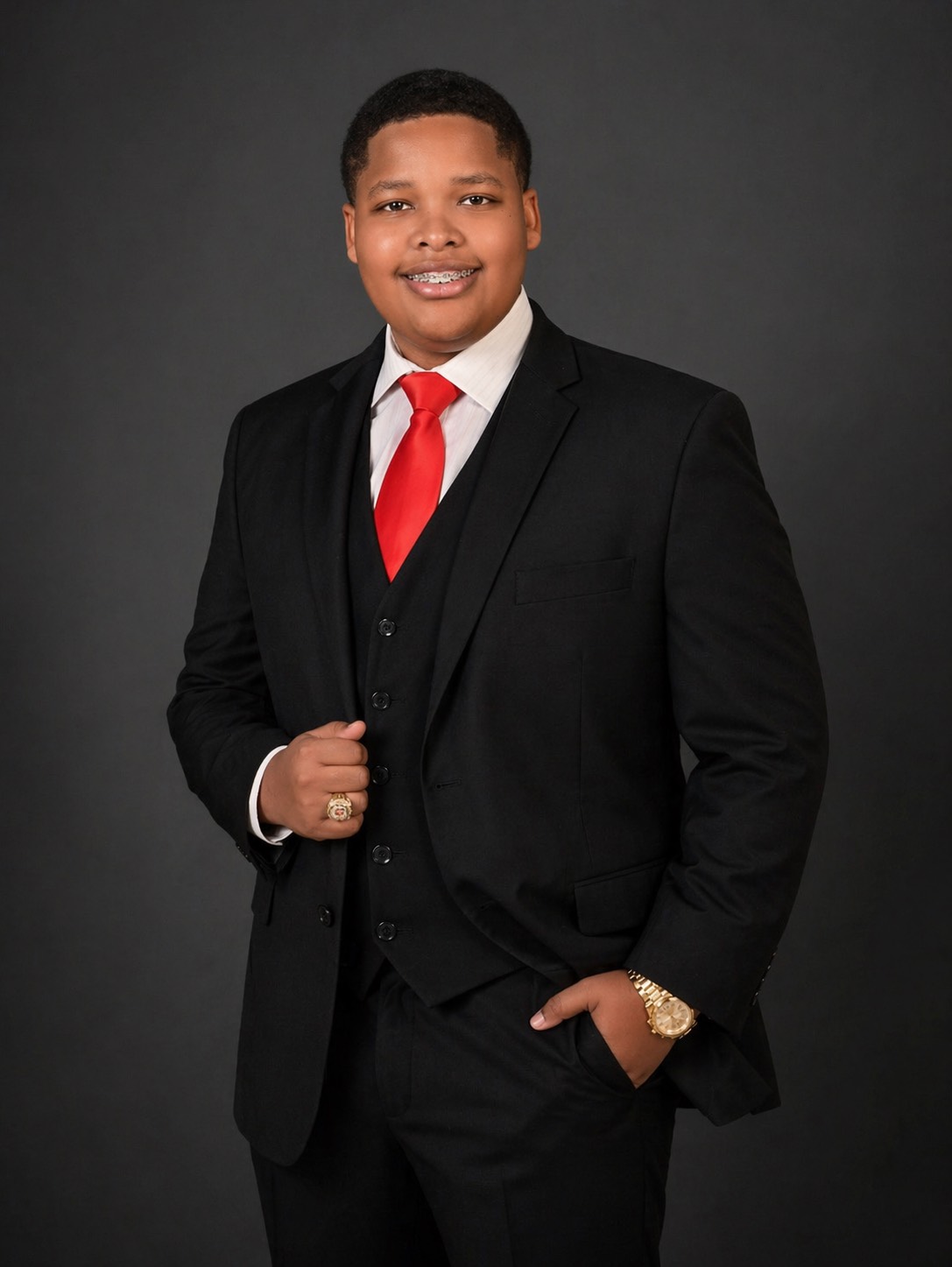 Young man in black suit, white shirt, red tie, smiling, standing against dark background.
