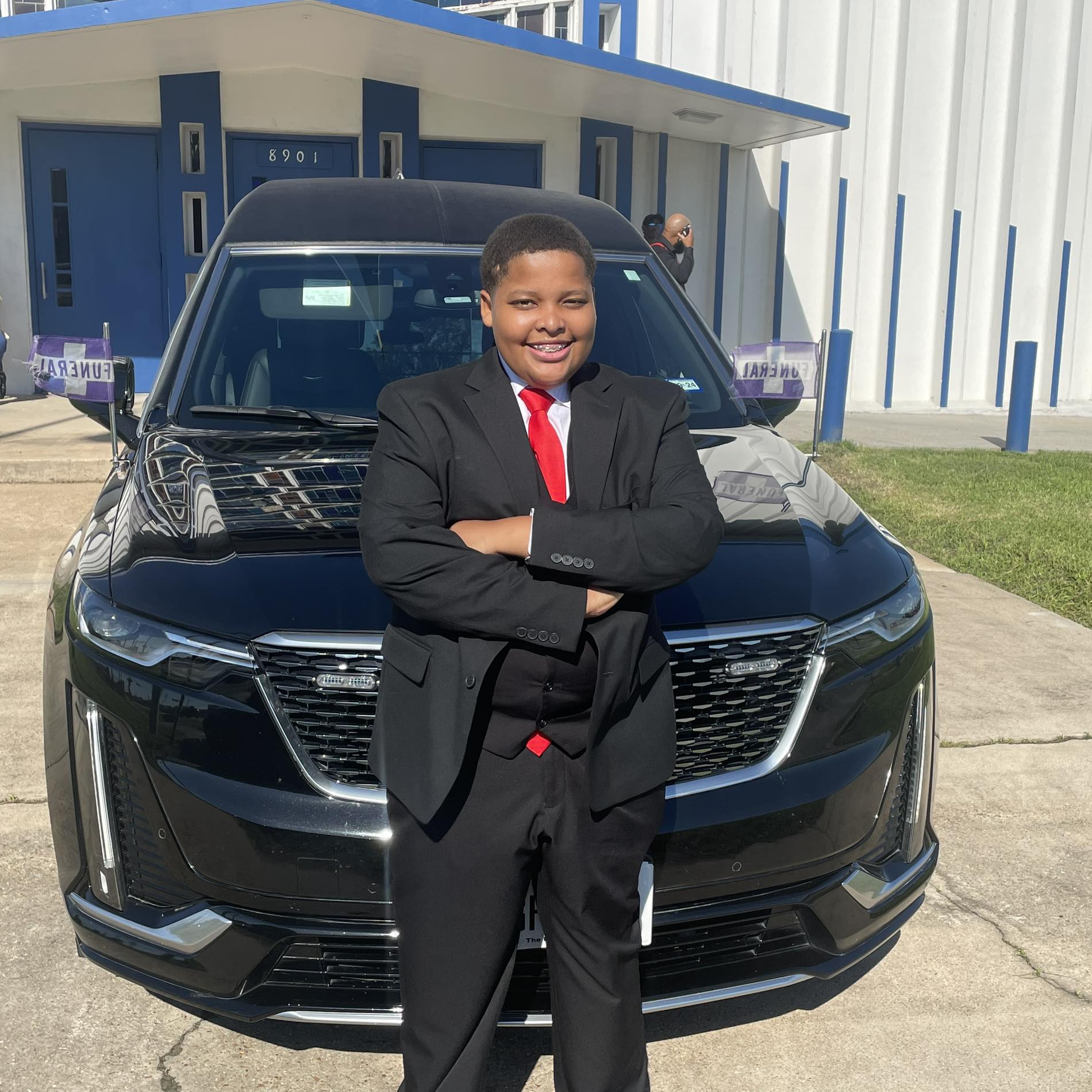 Young boy in a black suit with a red tie, smiling with arms crossed, standing in front of a black car.