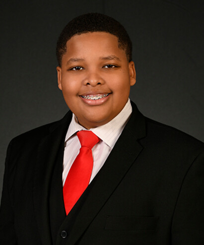 Young boy in a suit with a red tie, smiling, against a dark background.