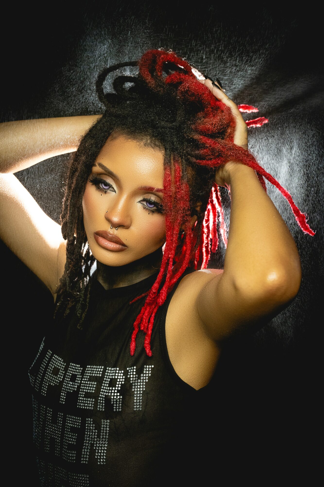 Woman with red and black dreadlocks, posing with arms raised, dark background, wearing a black sleeveless top.