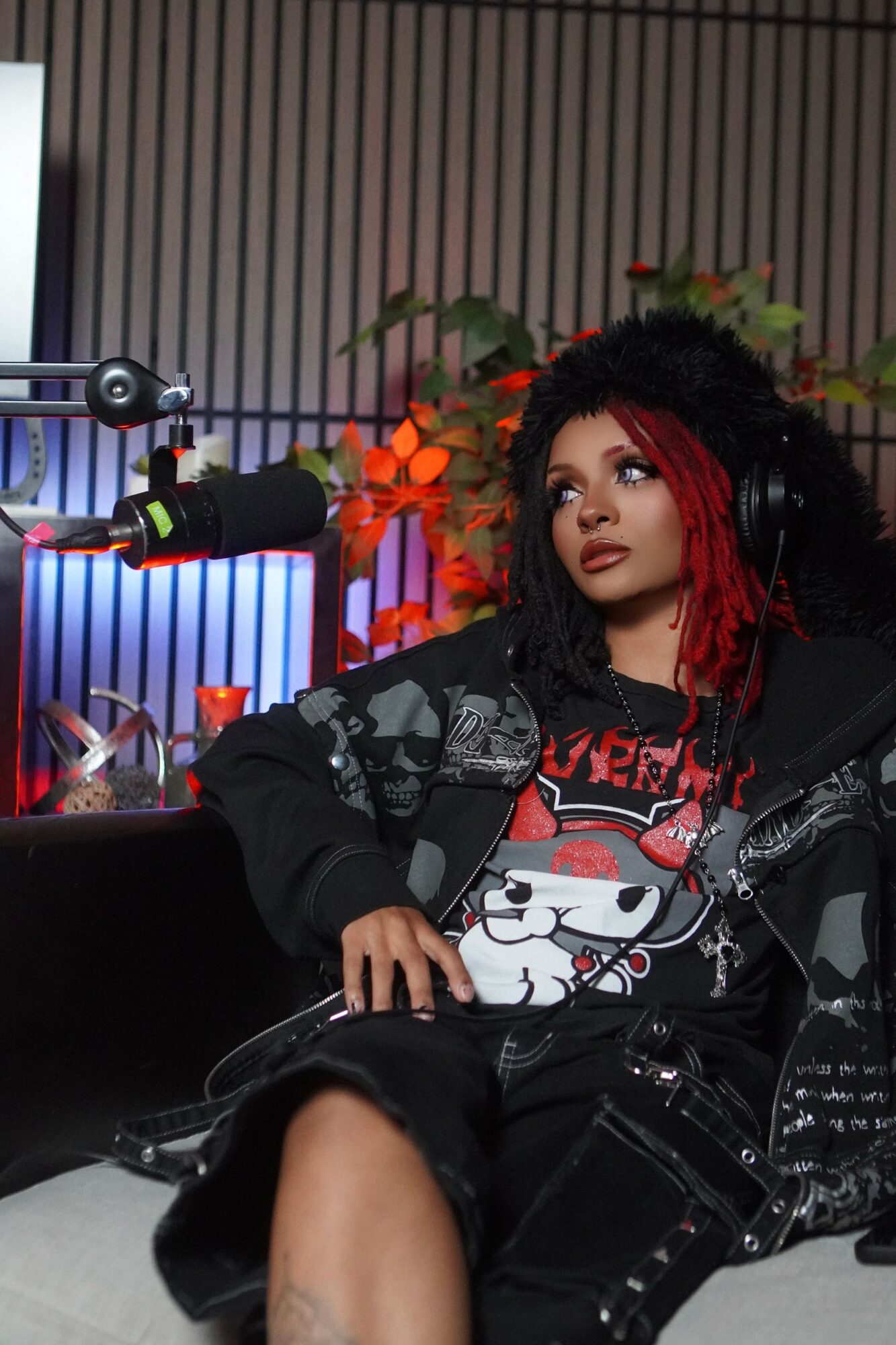 Woman with curly hair and red streaks wearing headphones, sitting near microphone with flowers in background.