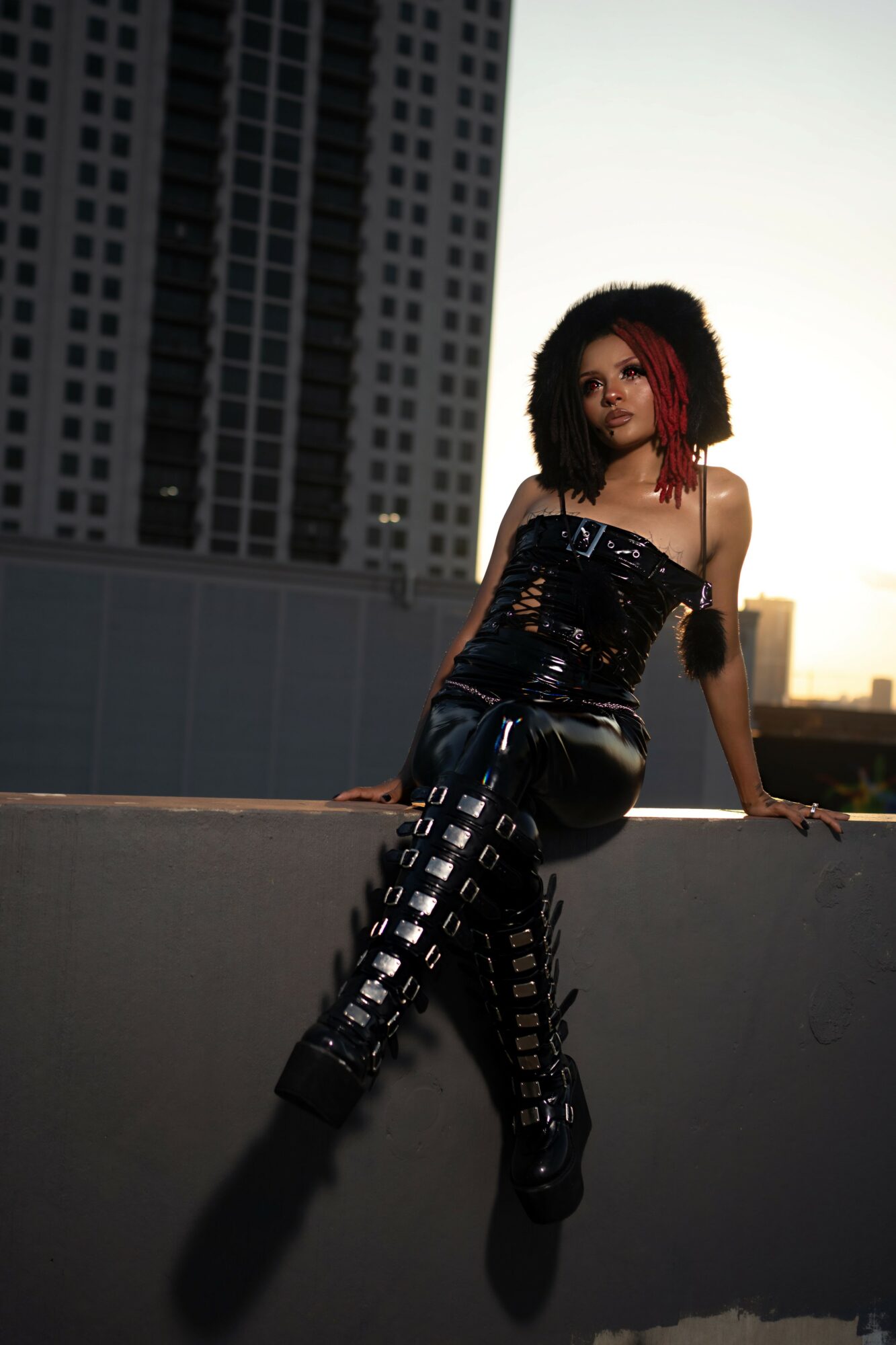Woman with curly hair in a black outfit sitting on a ledge in an urban setting during sunset.