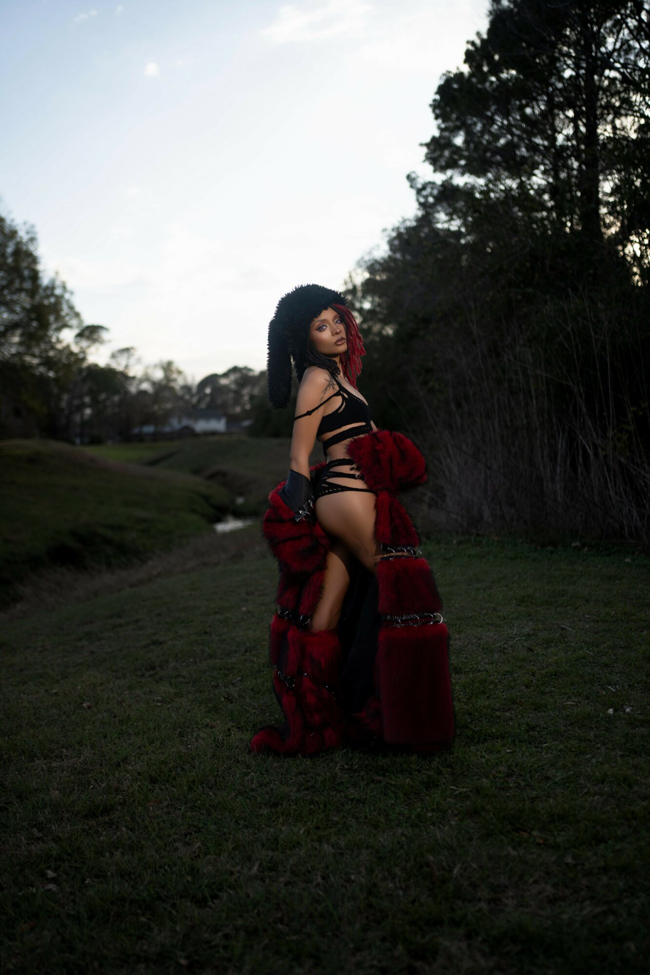 Person standing outdoors in a grassy area with trees, wearing black and red clothing, holding a red and black garment.
