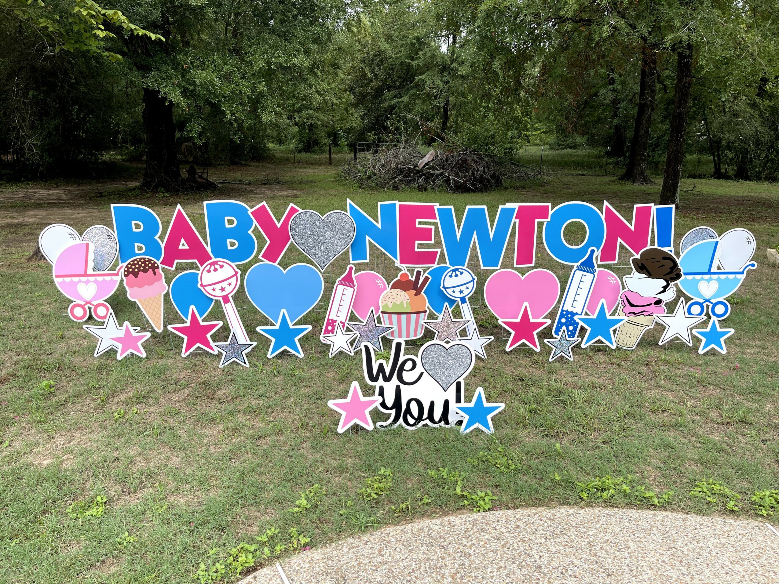 Decorative sign with colorful letters and icons celebrating a new baby, including hearts, stars, and strollers, outdoors on grass.