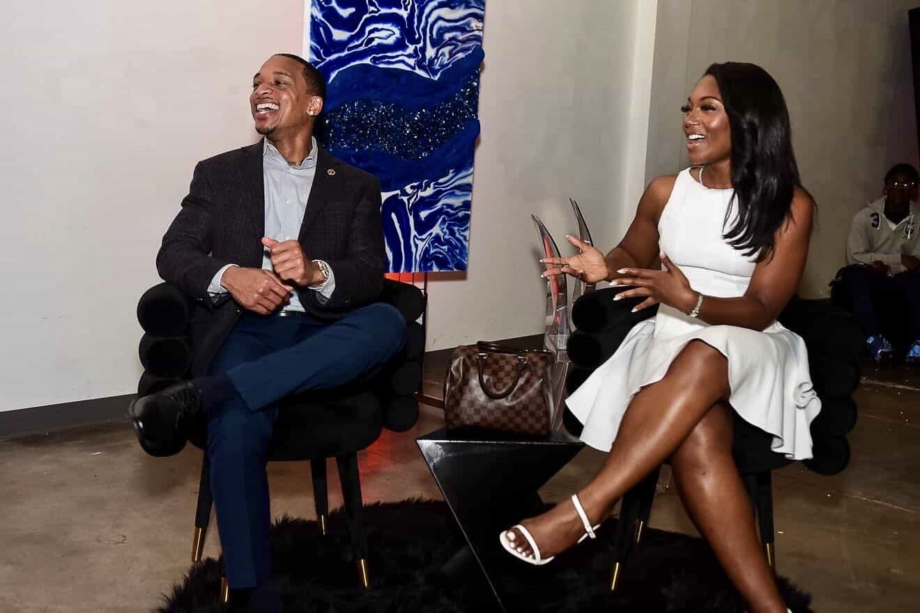 Two people sitting and smiling, one holding a phone, in an art gallery setting with artwork on the wall.