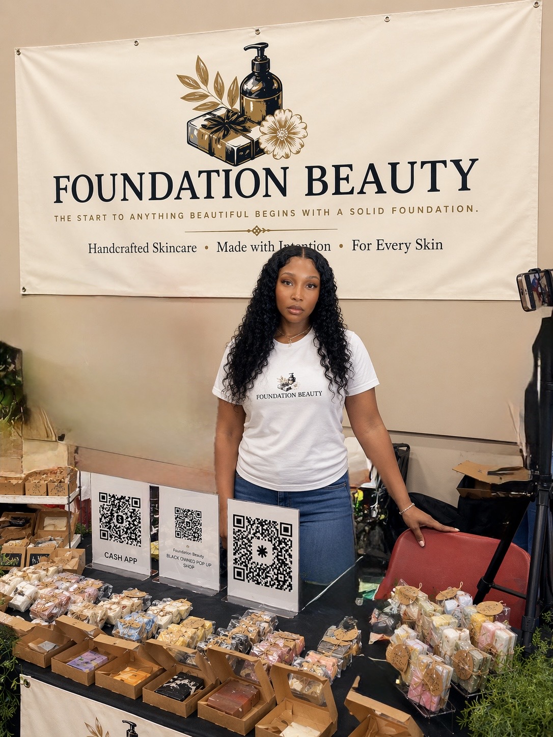 Woman standing behind a table with skincare products and QR codes, in front of a Foundation Beauty banner.