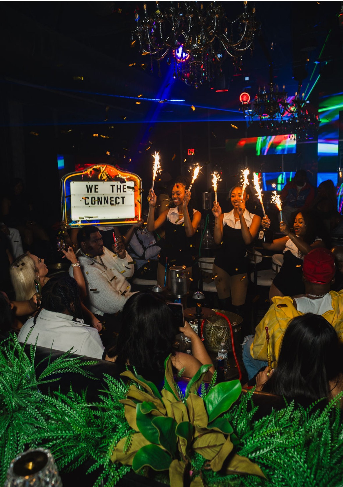 People in a nightclub with sparklers, a screen displaying 'WE THE CONNECT,' and colorful lights, with a crowd watching performers.