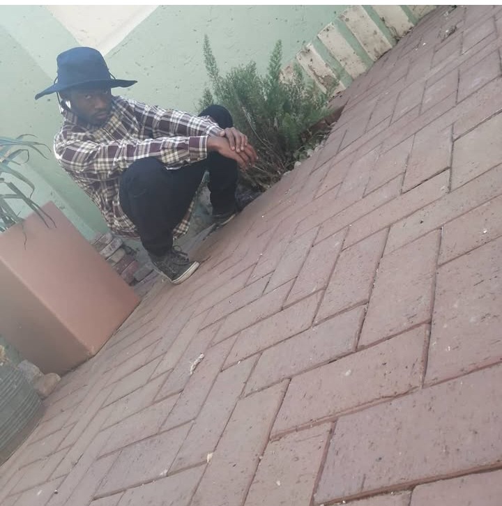 Person crouching next to a plant on a brick-paved surface, wearing a hat and plaid shirt.