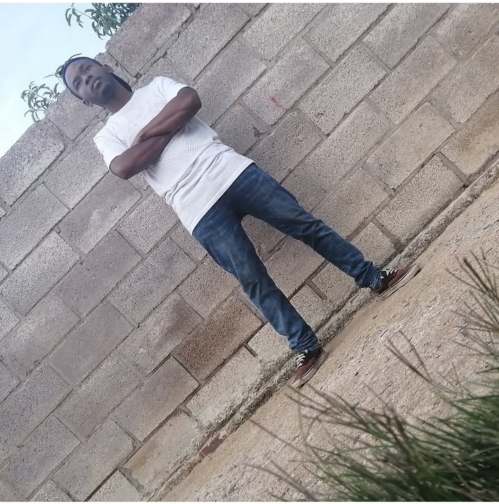 Person standing with arms crossed, leaning against a brick wall outdoors, wearing a white t-shirt and jeans.