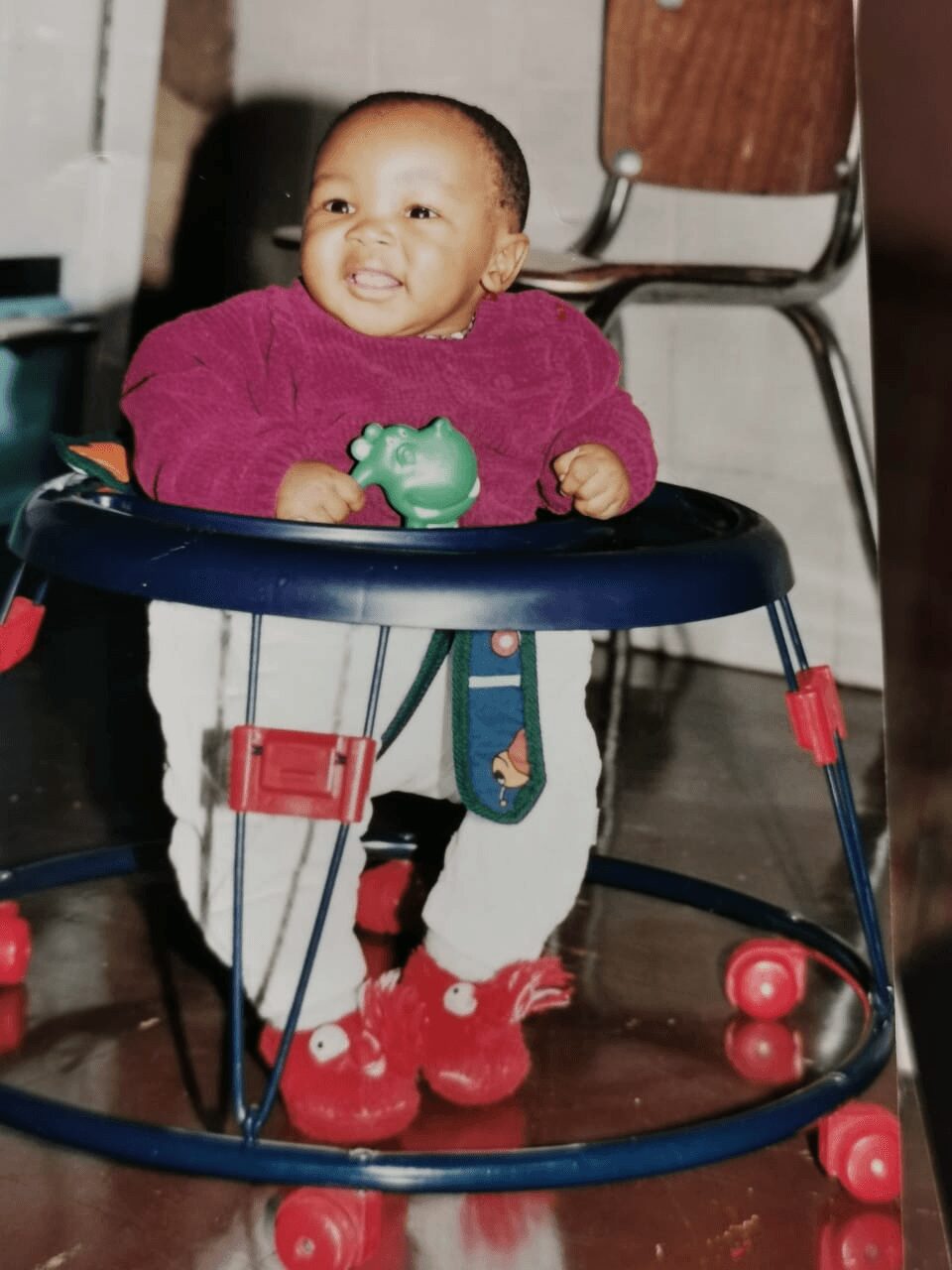 Baby in a walker smiling, holding a green toy, wearing a pink top and red shoes.