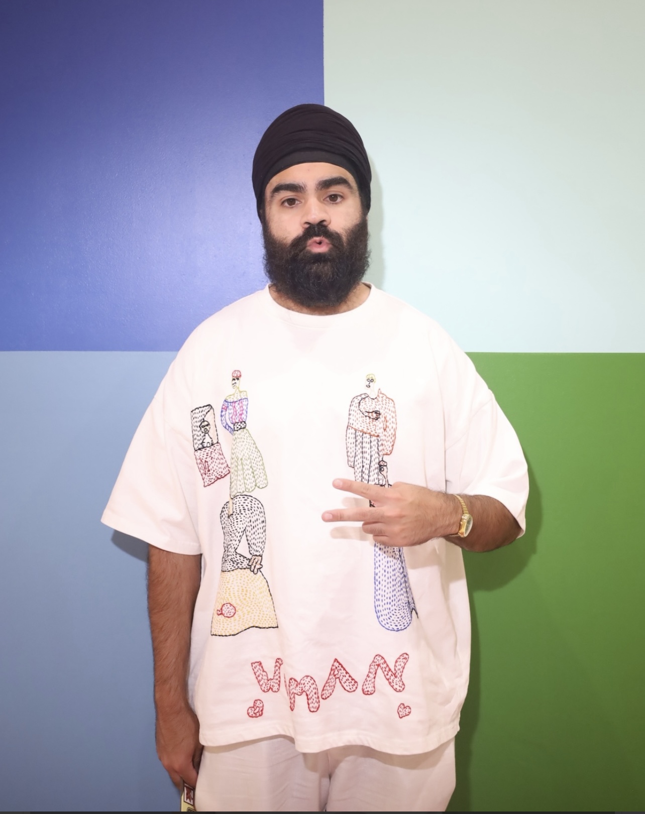 Man with a beard and turban standing against a colorful background, wearing a white oversized shirt with drawings.
