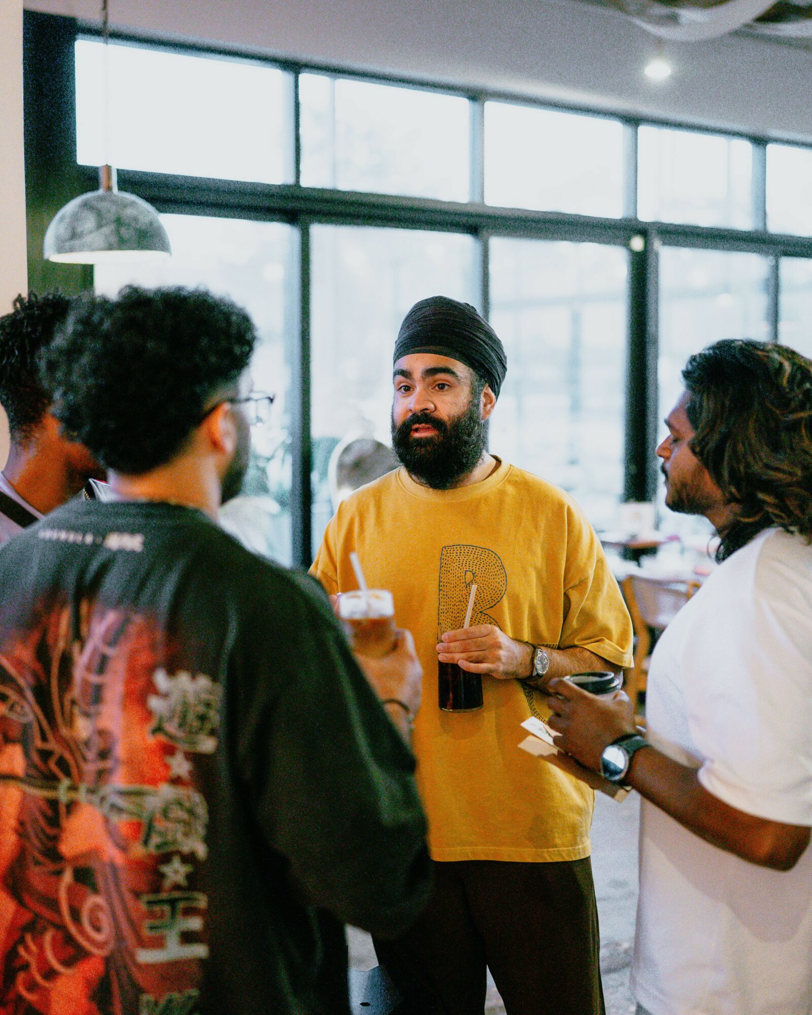Group of people talking indoors near large windows, one person holding a drink, casual setting.