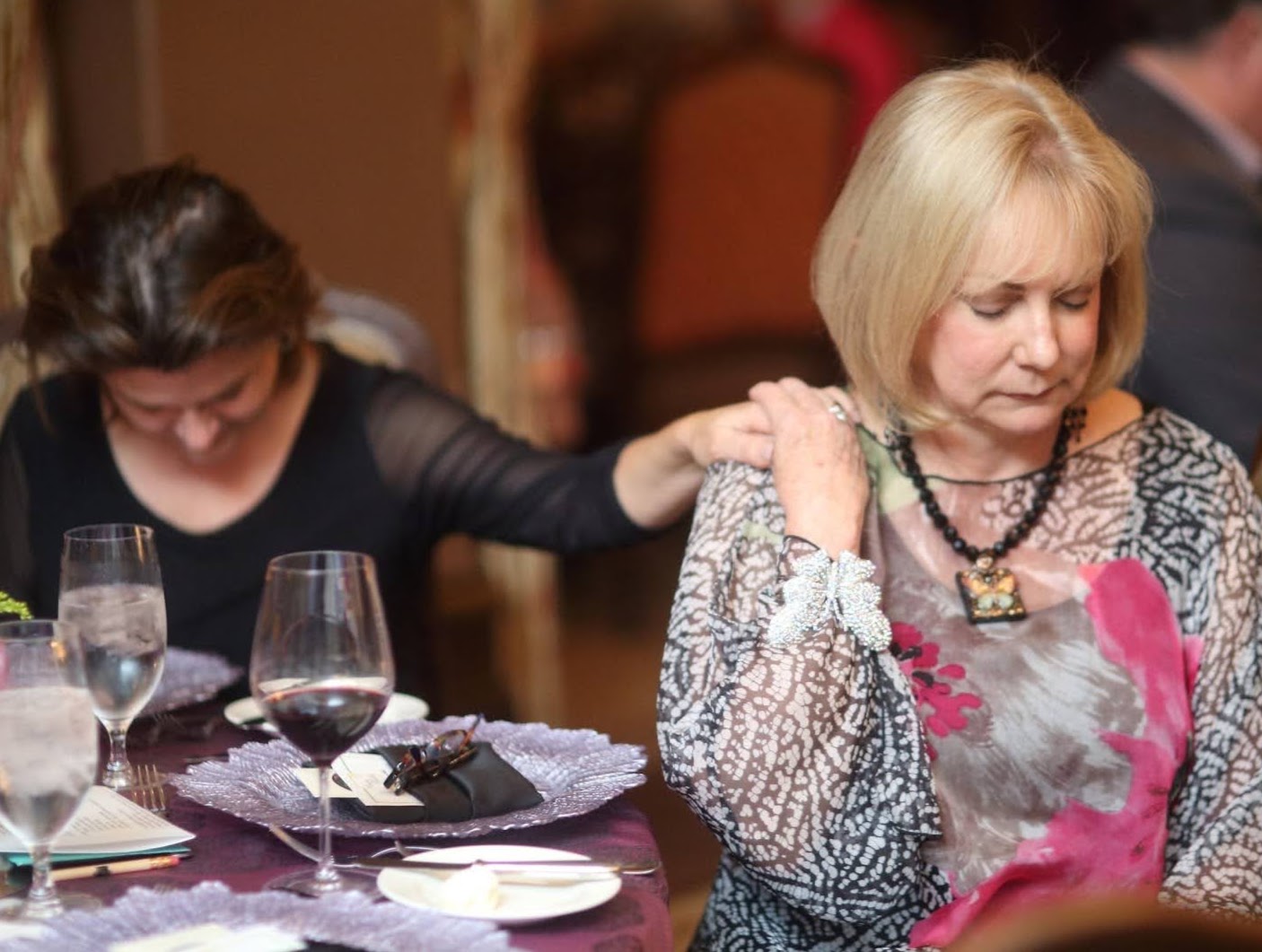 Two women at a table, one touching the other's shoulder, with glasses and tableware present.