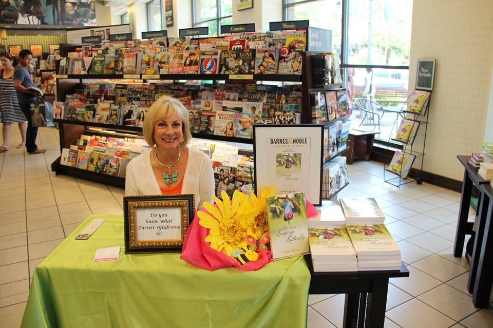 Woman with blonde hair sitting at a table with books and a bouquet of yellow flowers inside a bookstore.