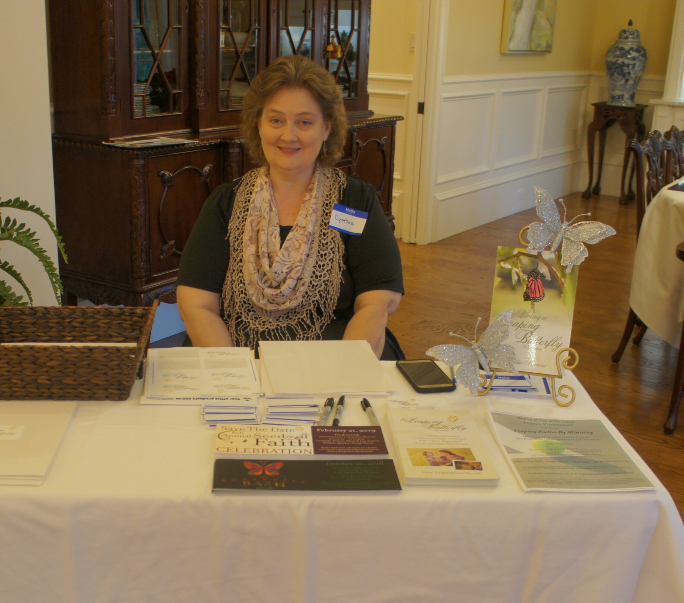 Woman sitting at a table with brochures and a basket, in a decorated room with wooden furniture and yellow walls.