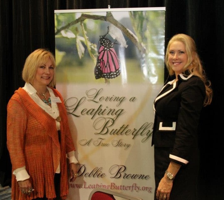 Two women stand beside a banner with a butterfly and text, smiling at the camera.