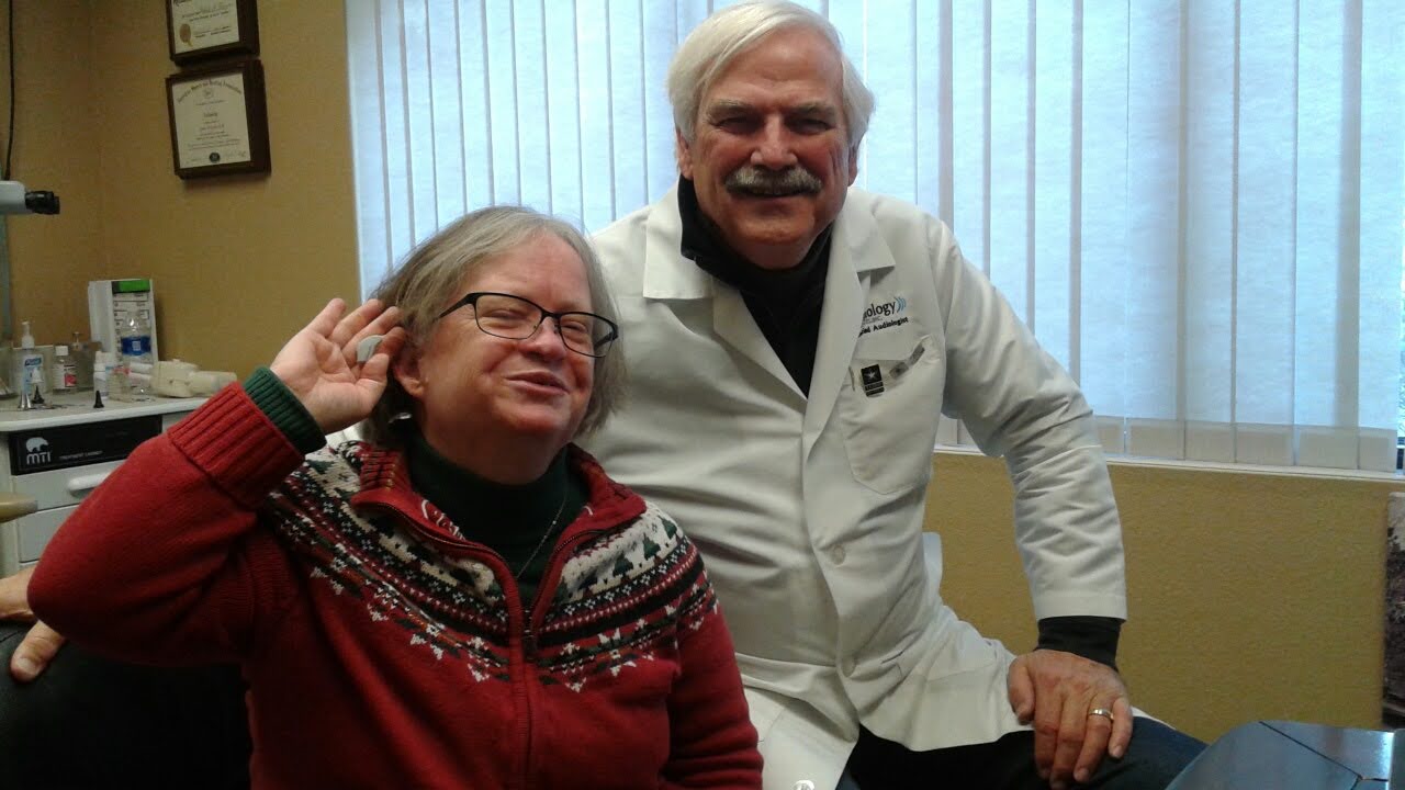 Two people, a woman and a man, smiling indoors with vertical blinds behind them.