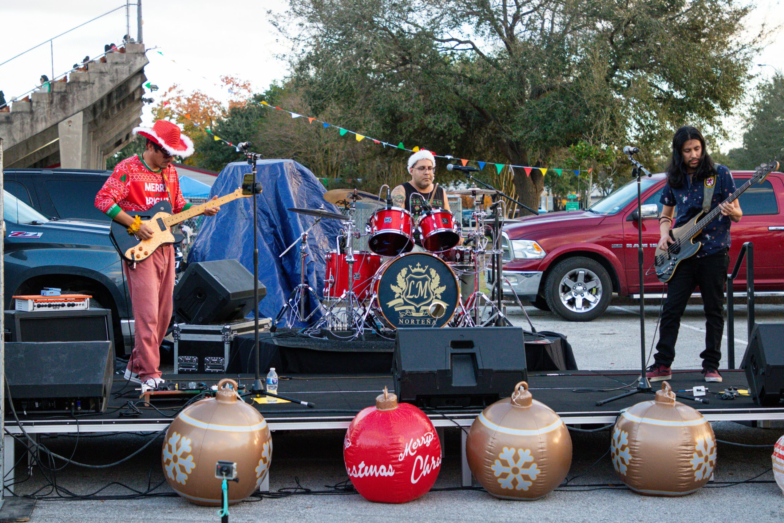 Three musicians perform on an outdoor stage with drums, guitars, and microphones, in a parking lot with trees and cars.