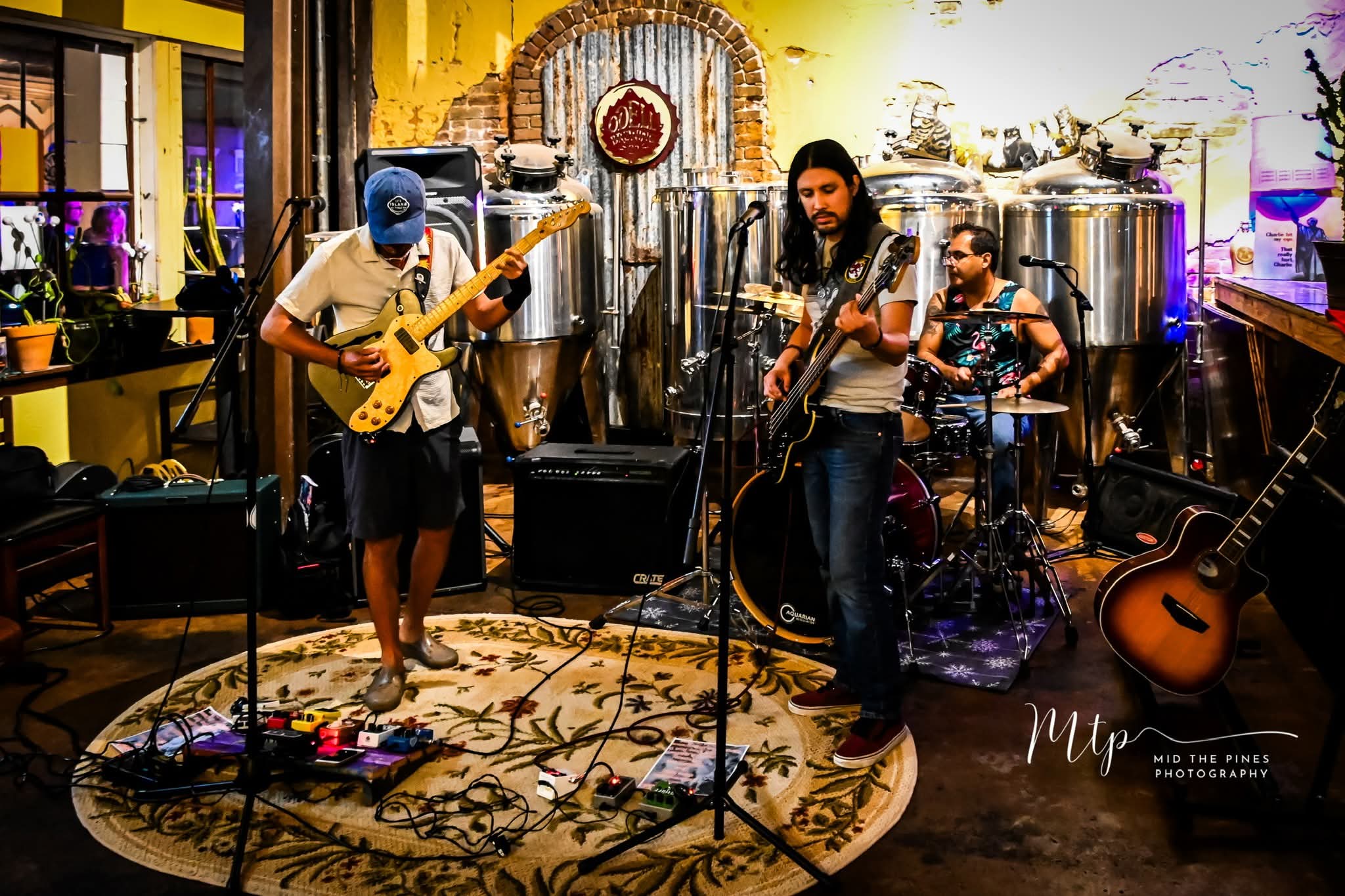 Three musicians perform with guitars and drums in a cozy, decorated indoor space with colorful lighting and musical equipment.