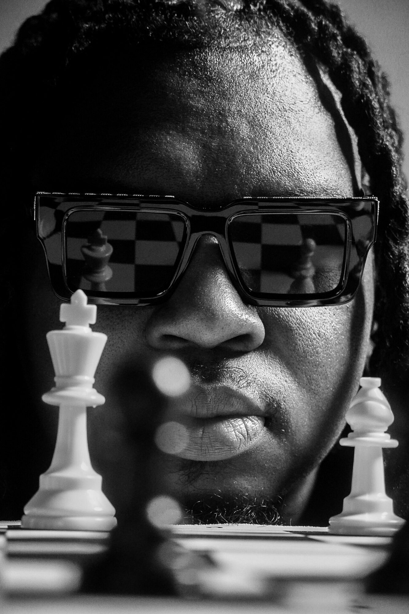 Close-up of a person with sunglasses playing chess, focus on face and chess pieces, black and white image.
