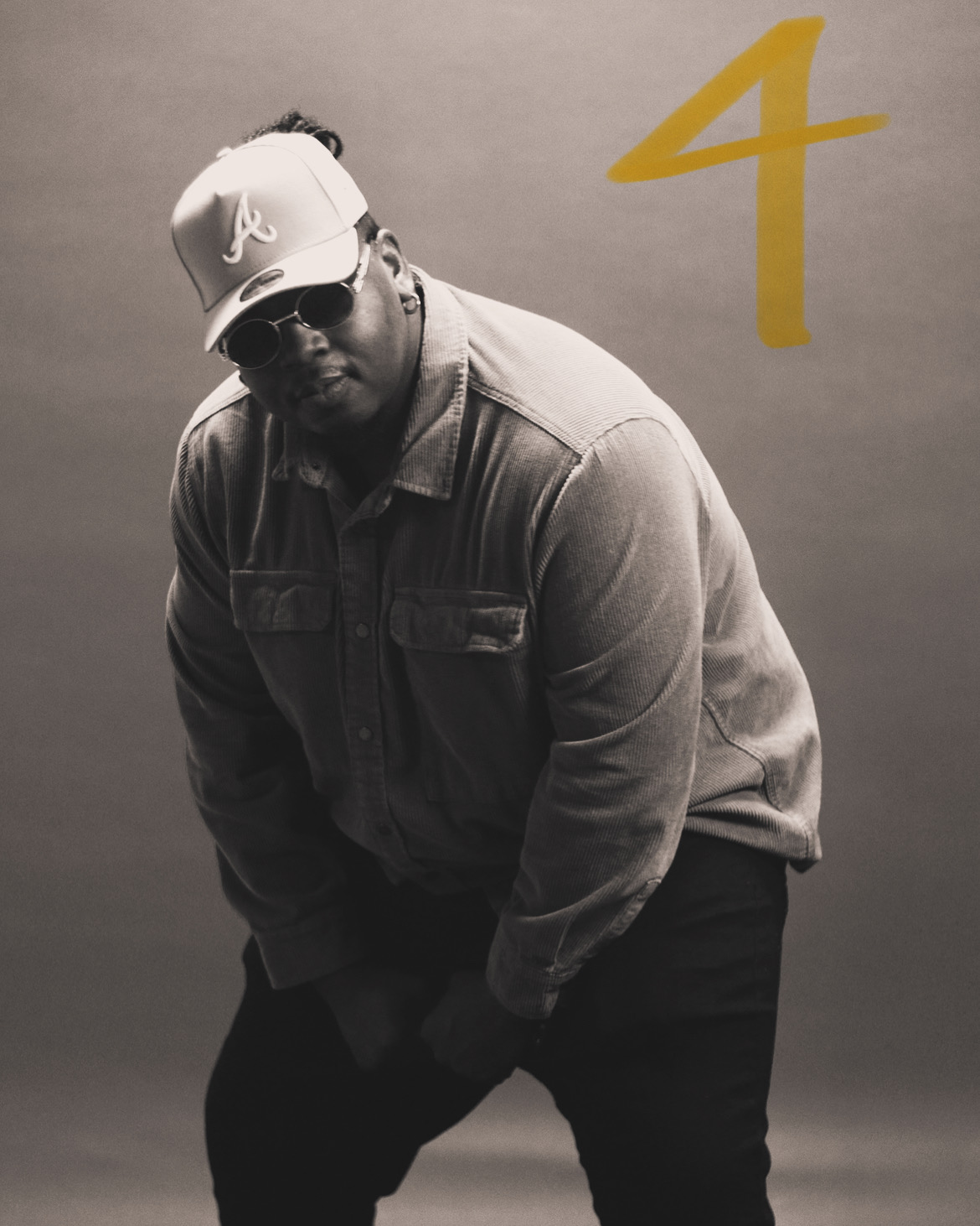Person wearing sunglasses, baseball cap, and denim jacket, standing against a plain background with a yellow number four.