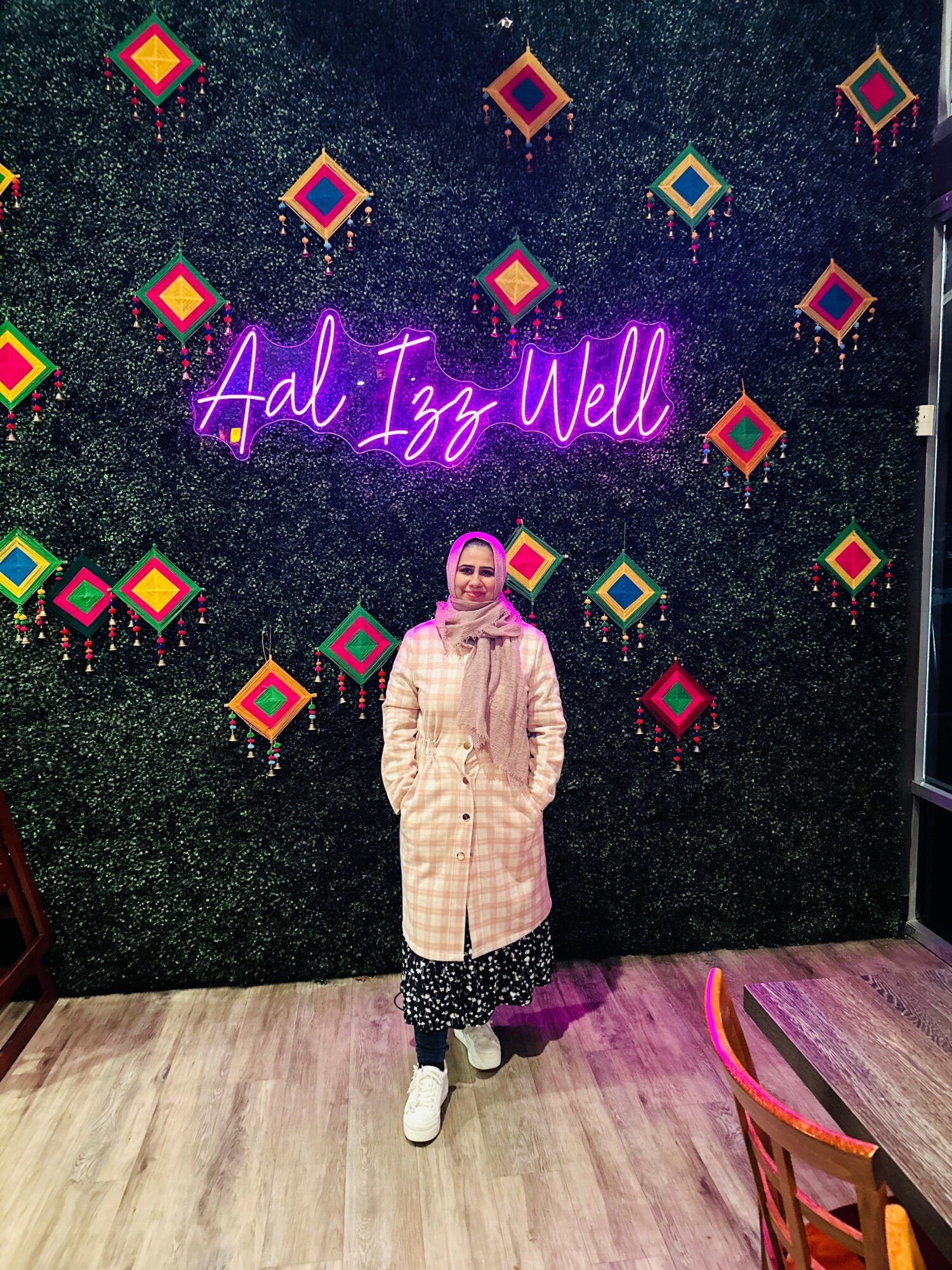 Person standing in front of a decorated wall with neon sign and colorful diamond shapes, wearing a coat and sneakers.