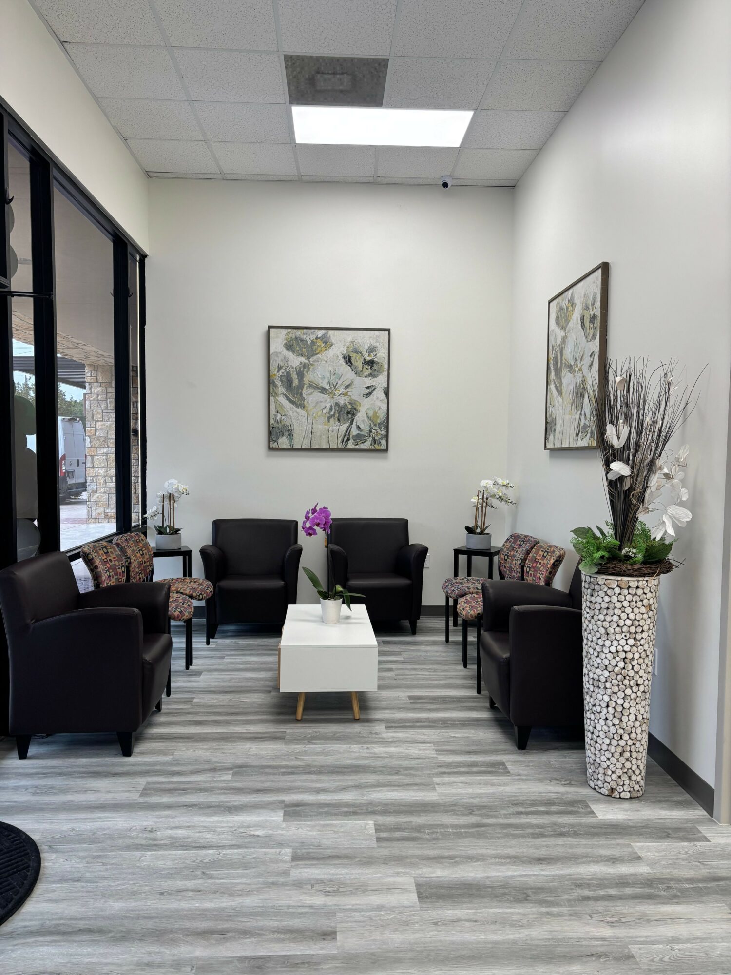 Waiting area with four black armchairs, a white coffee table, and a tall vase with flowers, against white walls.