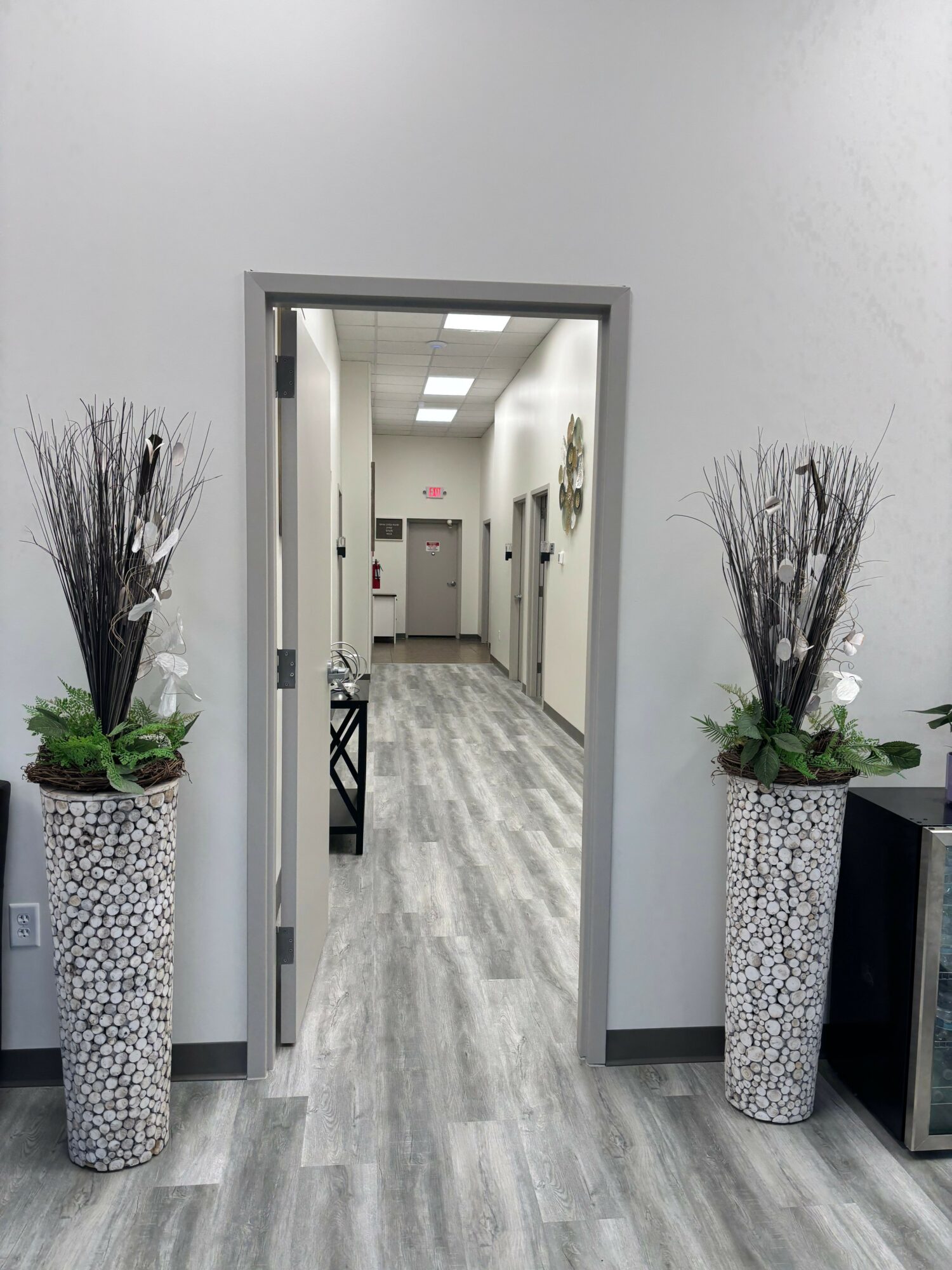 Hallway with gray flooring and white walls, two tall vases with plants on either side of doorway, and a bench inside.