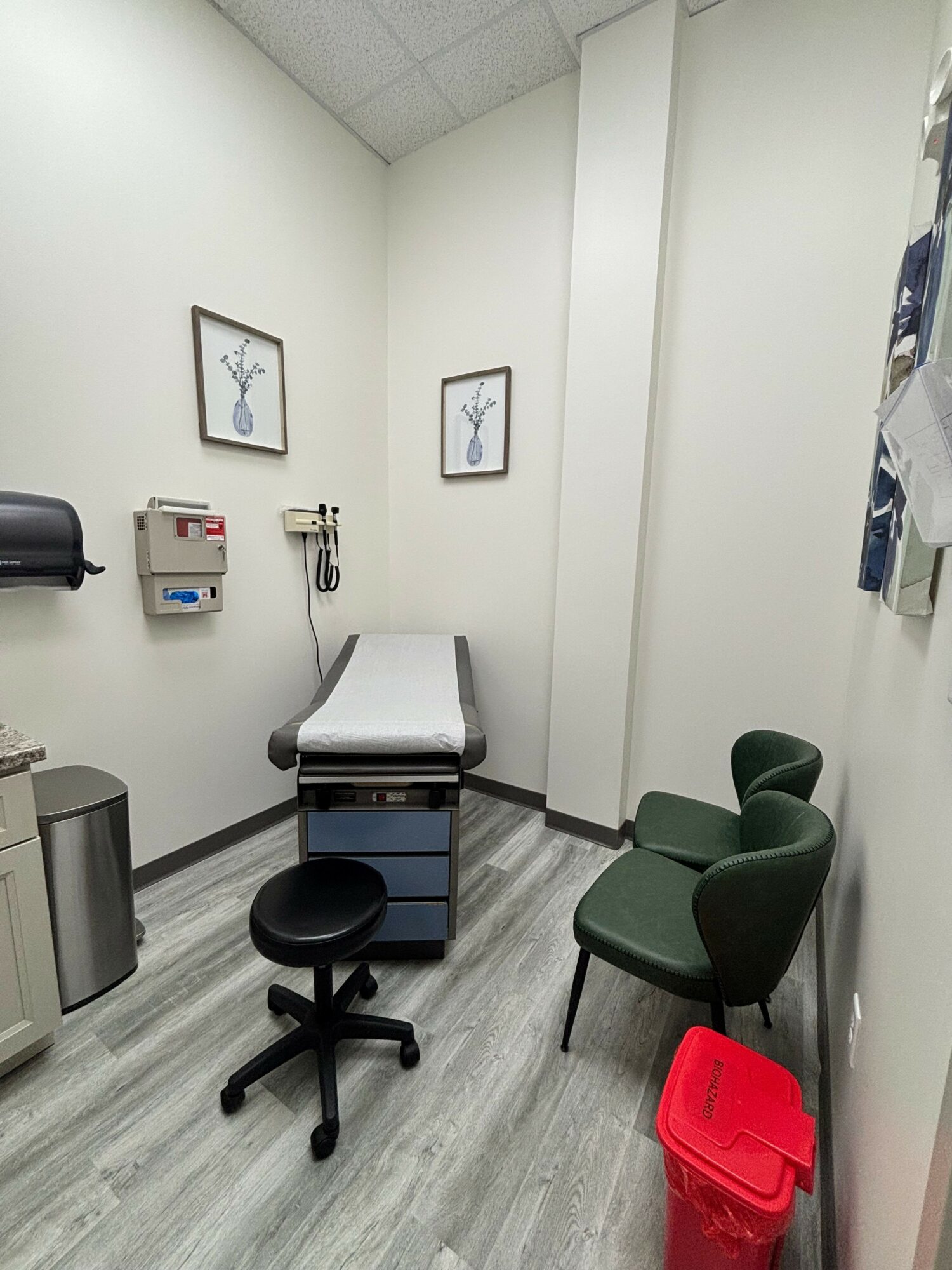 Small medical examination room with two chairs, a bed, and medical equipment on the wall.