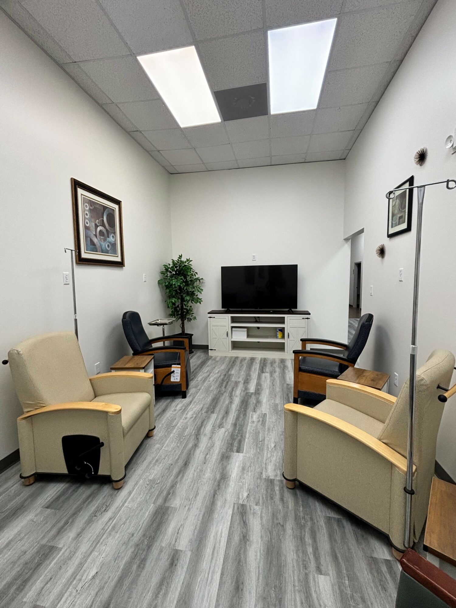 Small room with four beige armchairs, a TV on a white stand, a potted plant, and framed pictures on white walls.