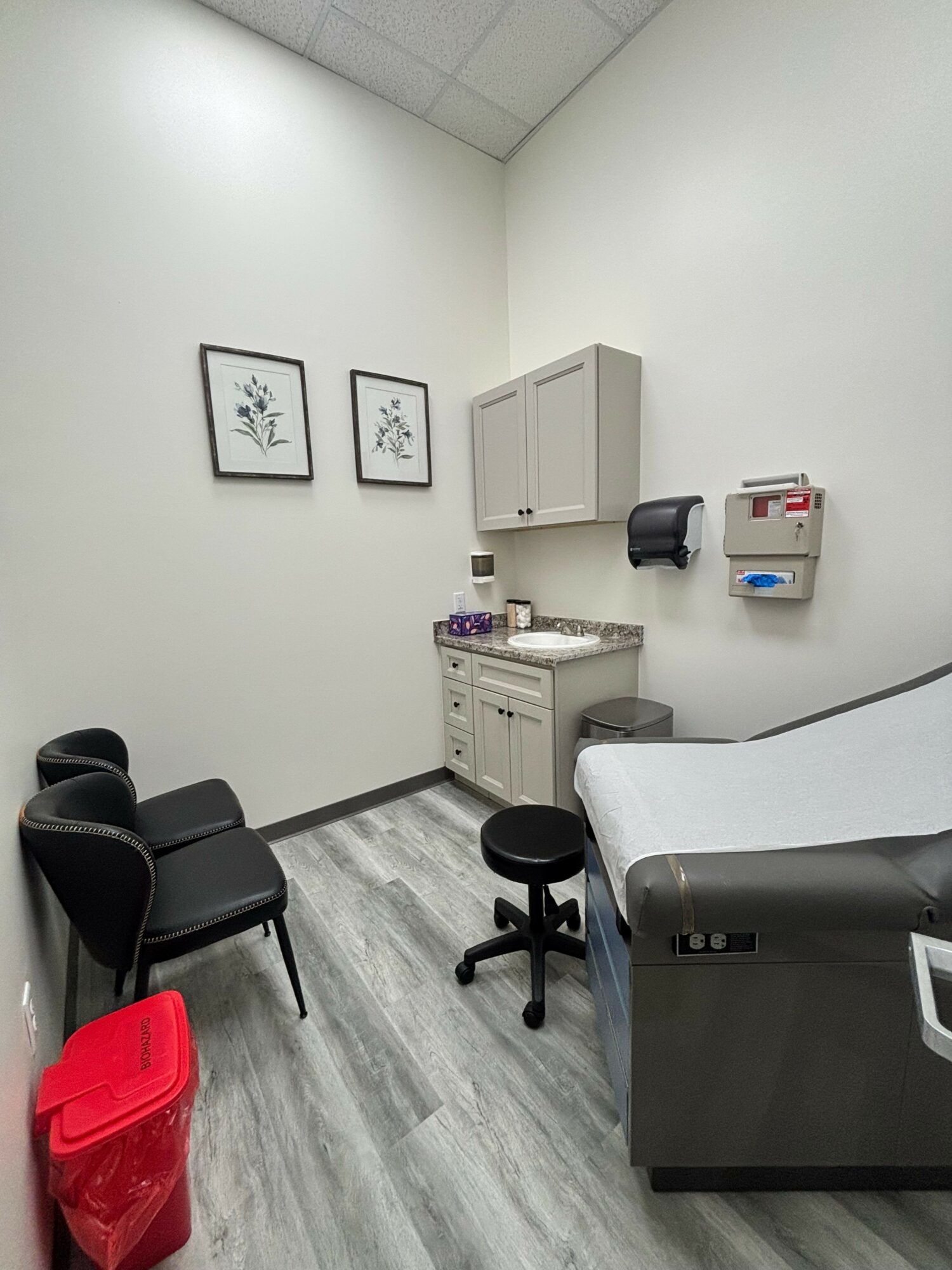 Medical examination room with chairs, cabinet, examination bed, and wall-mounted supplies, in neutral colors and wood flooring.