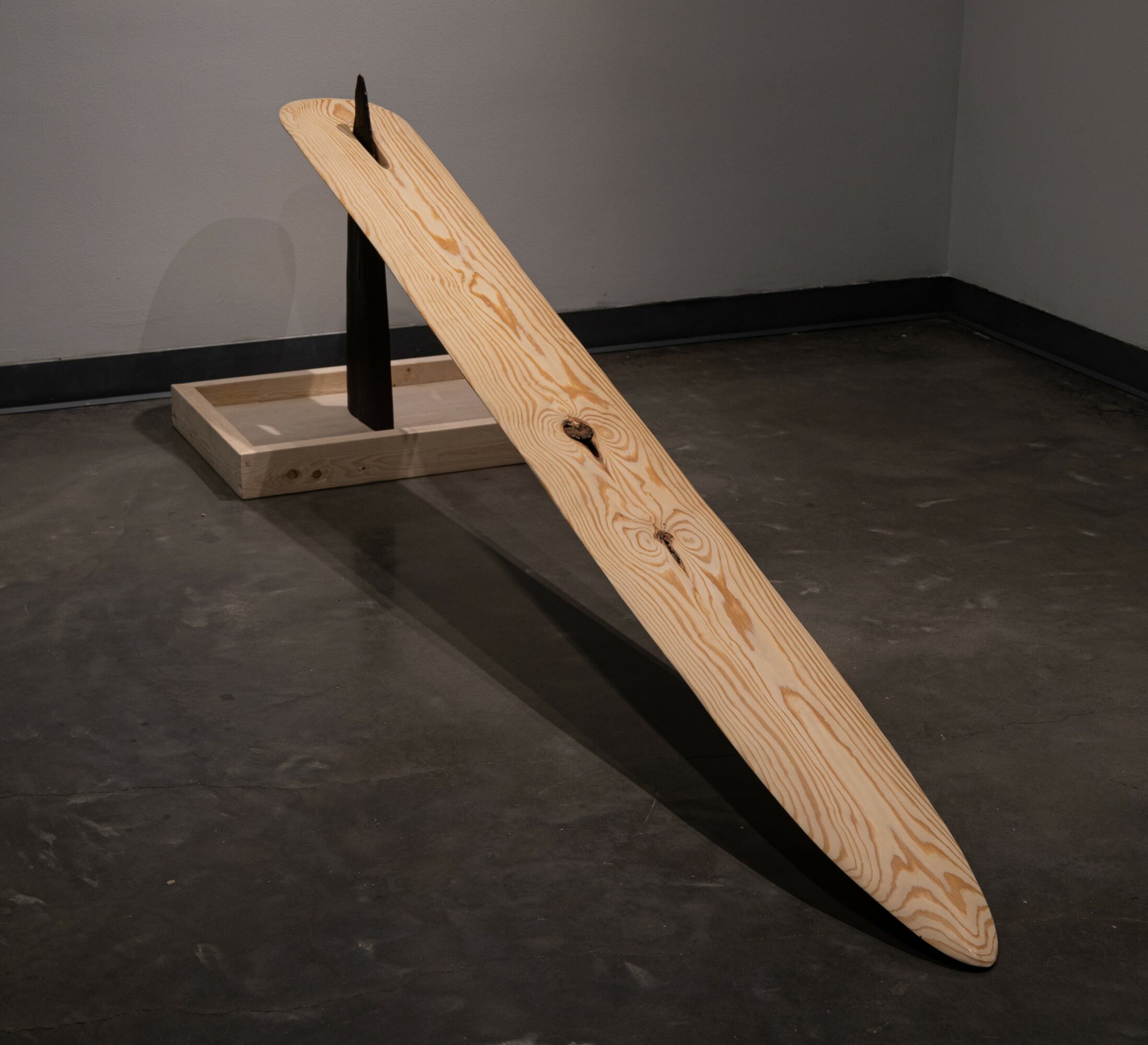 Wooden surfboard leaning against a black stand on a dark floor in a room with gray walls.