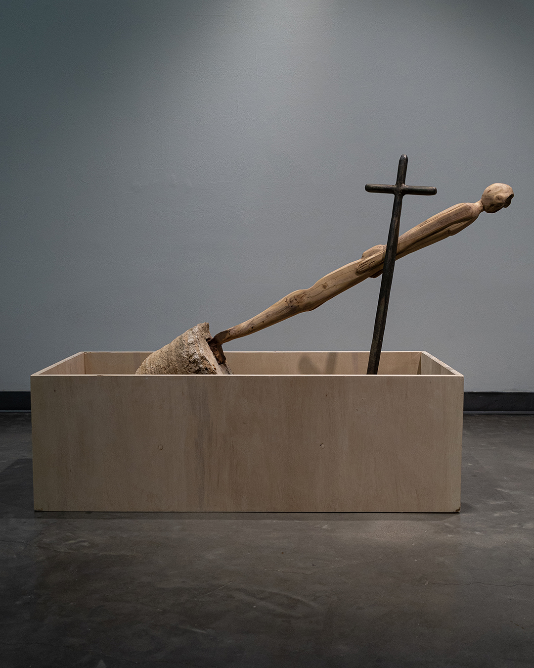 Sculpture of a wooden cross and a twisted wooden arm in a rectangular container, against a plain background.
