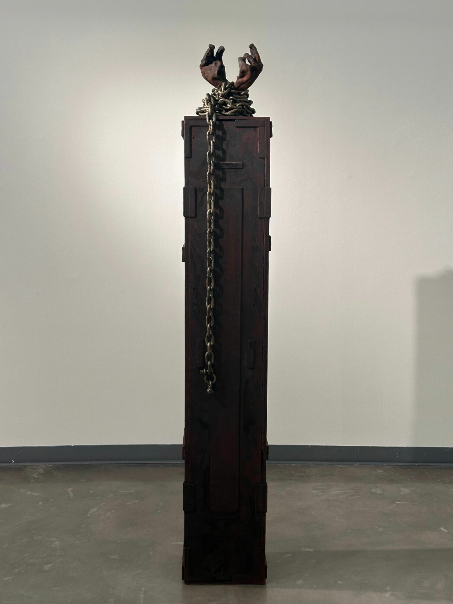 Tall dark wooden pedestal with a chain and a decorative object on top, displayed in a gallery setting.