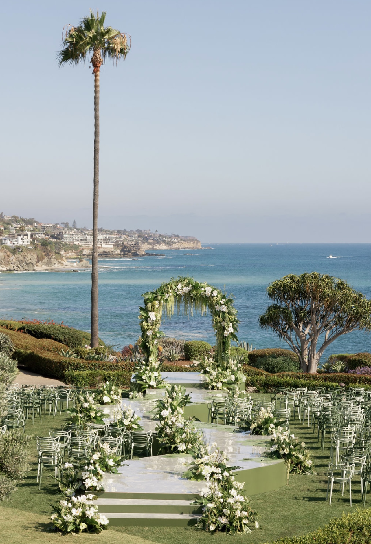 Outdoor wedding setup with floral arch, chairs, and ocean view in the background, including a tall palm tree and a tree on the right.