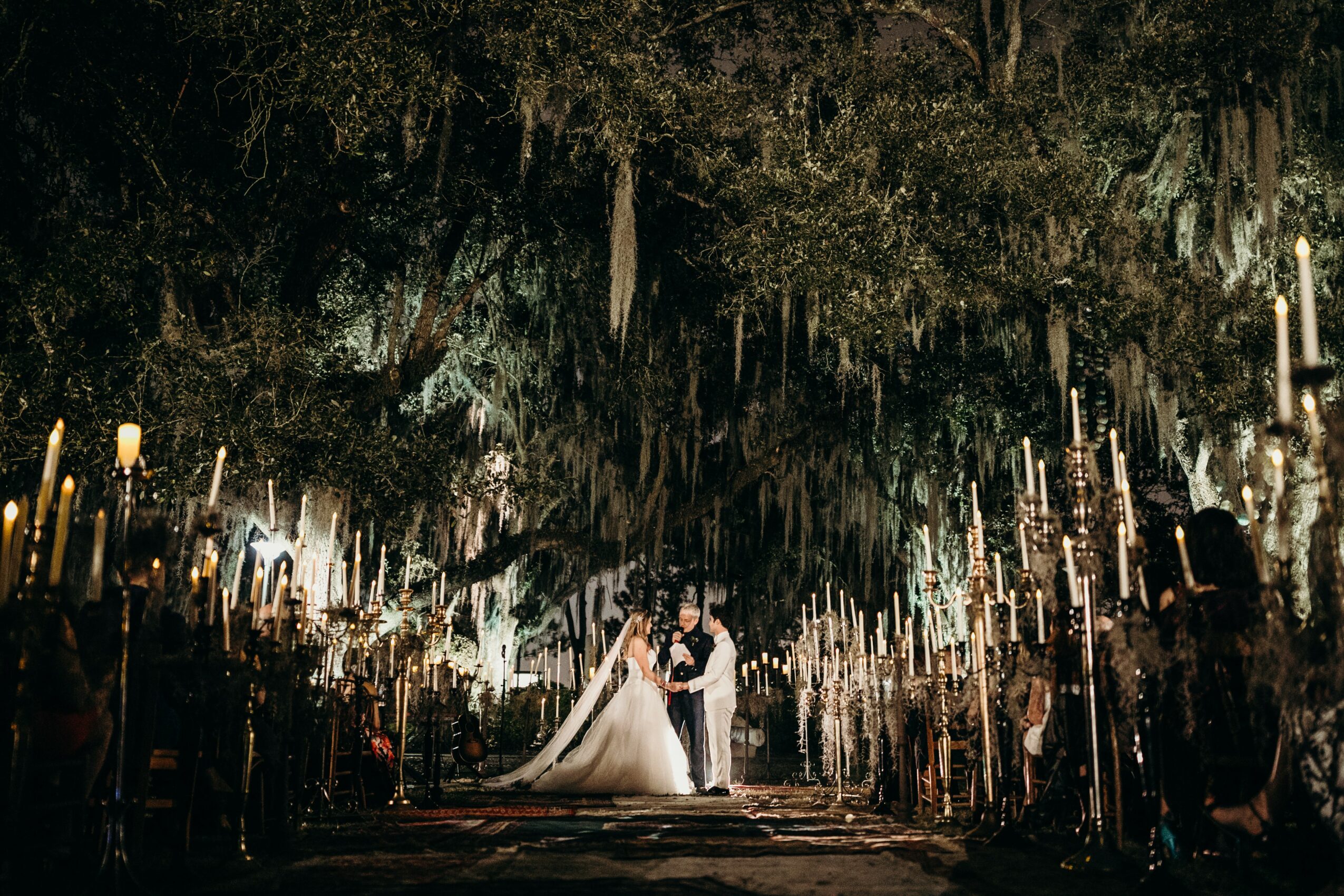 A bride and groom stand in a decorated outdoor setting at night with candles and trees around them.