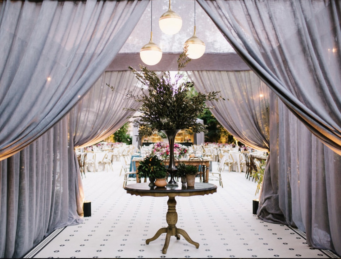 Elegant event space with draped curtains, a central table with a tree and flowers, and hanging lights.