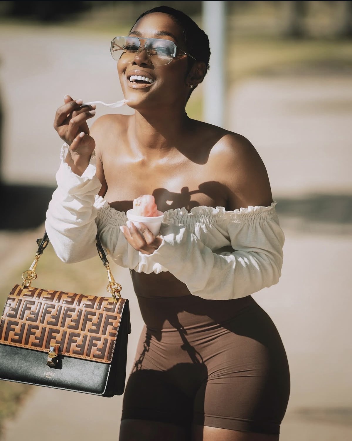Smiling woman with sunglasses holding ice cream cone outdoors, carrying a patterned handbag, wearing off-shoulder top and brown shorts.