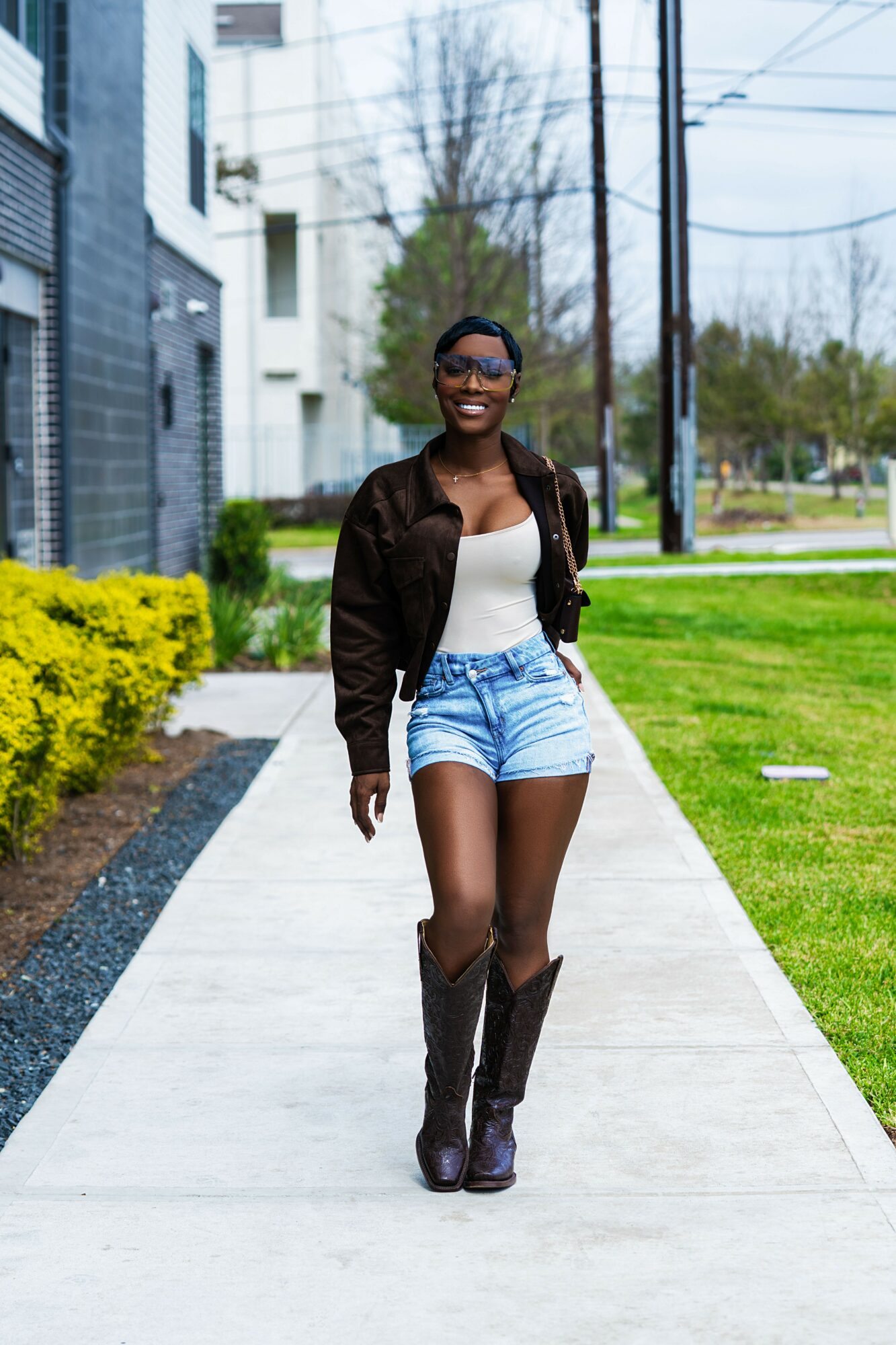 Young woman walking on sidewalk, smiling, wearing sunglasses, white top, denim shorts, and knee-high boots.