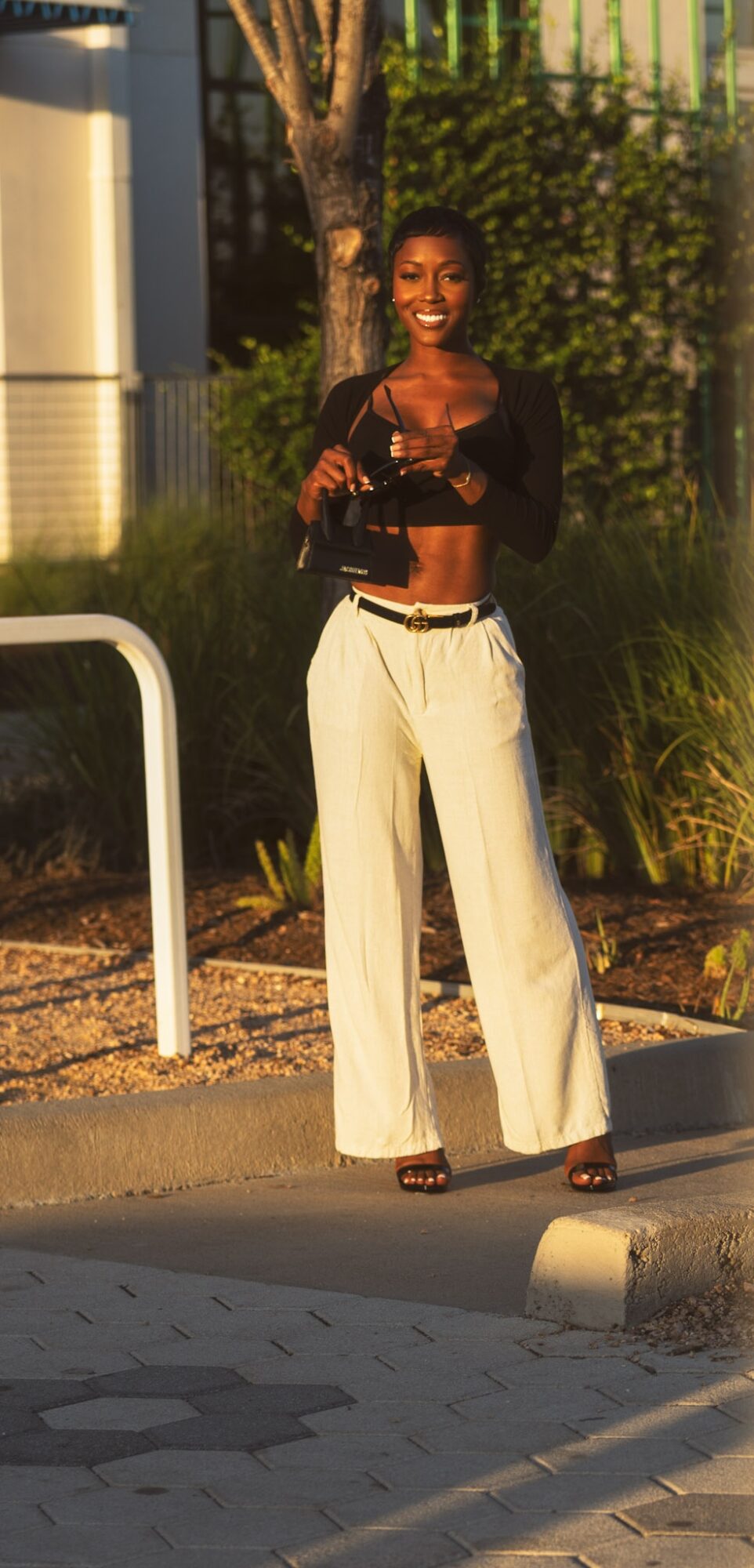 Woman standing outdoors, smiling, wearing black top and white wide-leg pants, with trees and a fence in background.