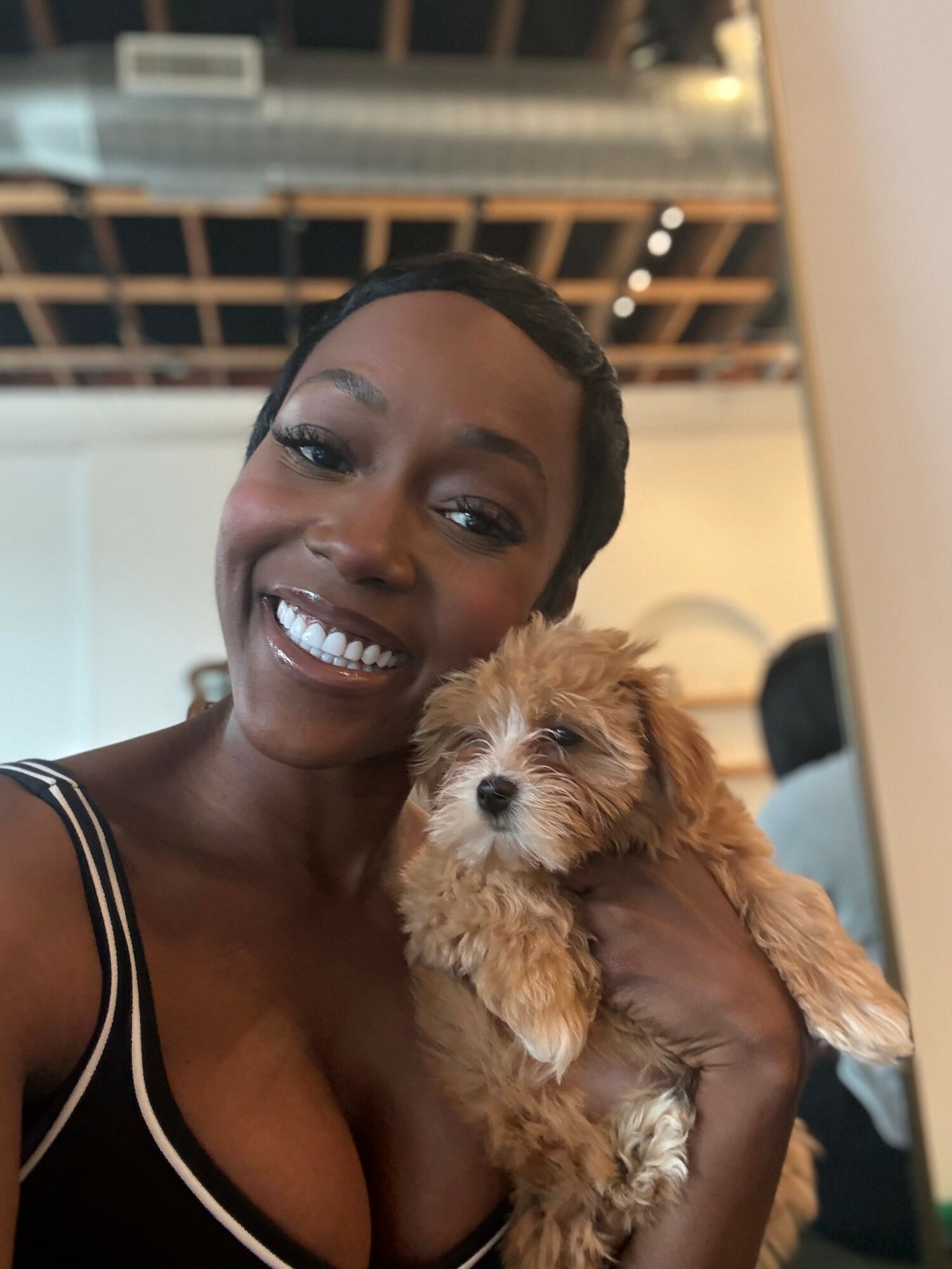 Smiling woman holding a small, fluffy puppy indoors with a ceiling and lights visible above.