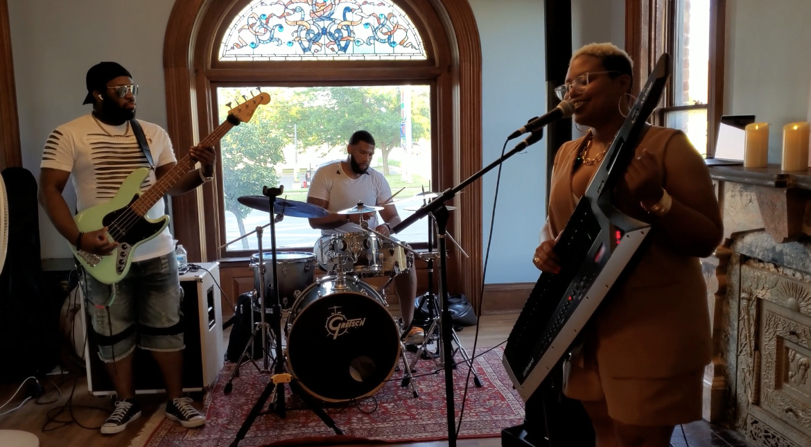 Three musicians performing indoors with a window and fireplace in the background.