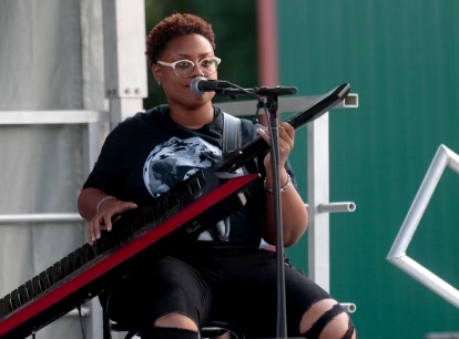 Person playing keyboard and singing into microphone outdoors, wearing glasses and a black shirt.