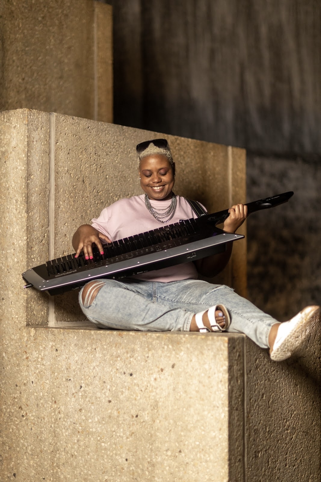 Person sitting on concrete steps, smiling, holding a laptop, wearing a pink shirt, jeans, and white shoes.