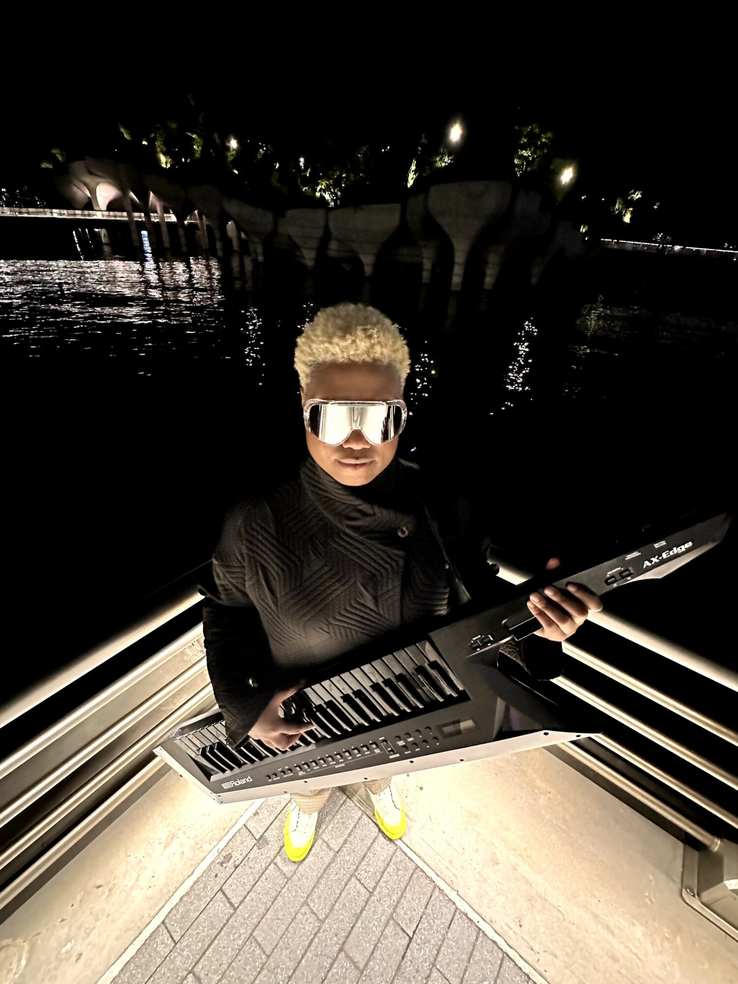 Person with short curly hair wearing sunglasses, holding a keyboard, standing on a bridge at night with water and lights in background.